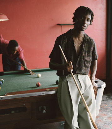 A man with a cue stick leans against a pool table while another man takes a shot